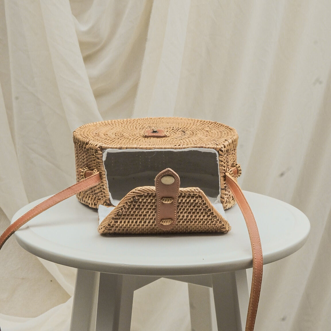 Woven handbag with leather strap on a small round table against a white curtain background