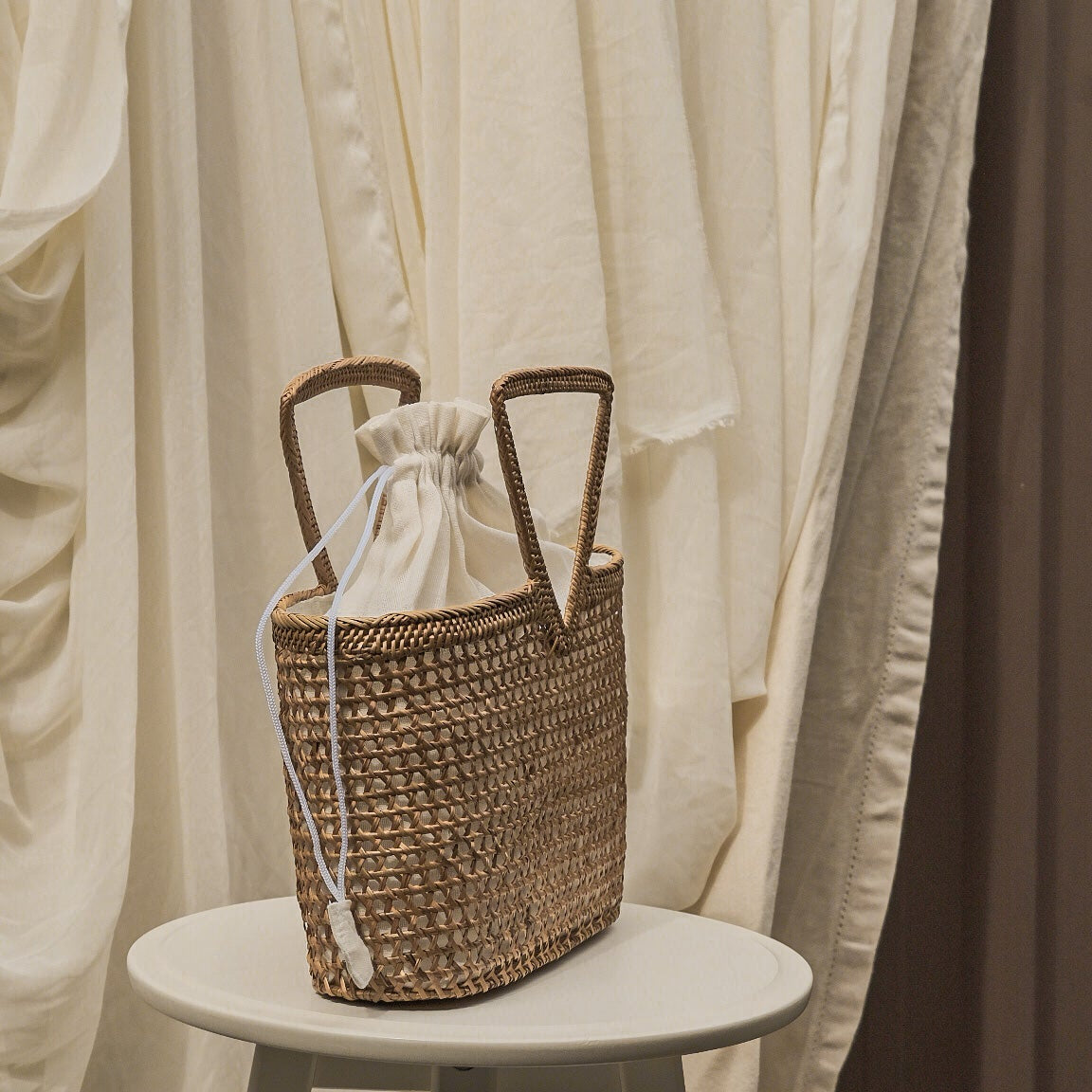 Woven basket with a sheer fabric drawstring on a white stool against a draped curtain background