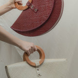 Two half-circle handbags with wooden handles held by a person against a neutral background.