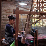 A woman artisan weaving a handloom blanket using a traditional wooden loom in a rustic workshop. She carefully threads the fabric, showcasing the intricate craftsmanship involved in creating handwoven textiles. Natural light filters through the brick lattice wall, highlighting the artisan’s dedication to preserving traditional weaving techniques.