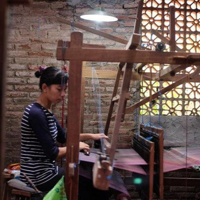 A woman artisan weaving a handloom blanket using a traditional wooden loom in a rustic workshop. She carefully threads the fabric, showcasing the intricate craftsmanship involved in creating handwoven textiles. Natural light filters through the brick lattice wall, highlighting the artisan’s dedication to preserving traditional weaving techniques.