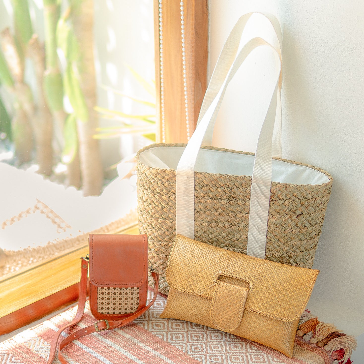 Woven bags including a large tote, small crossbody, and clutch on a textured surface with a blurred natural background.