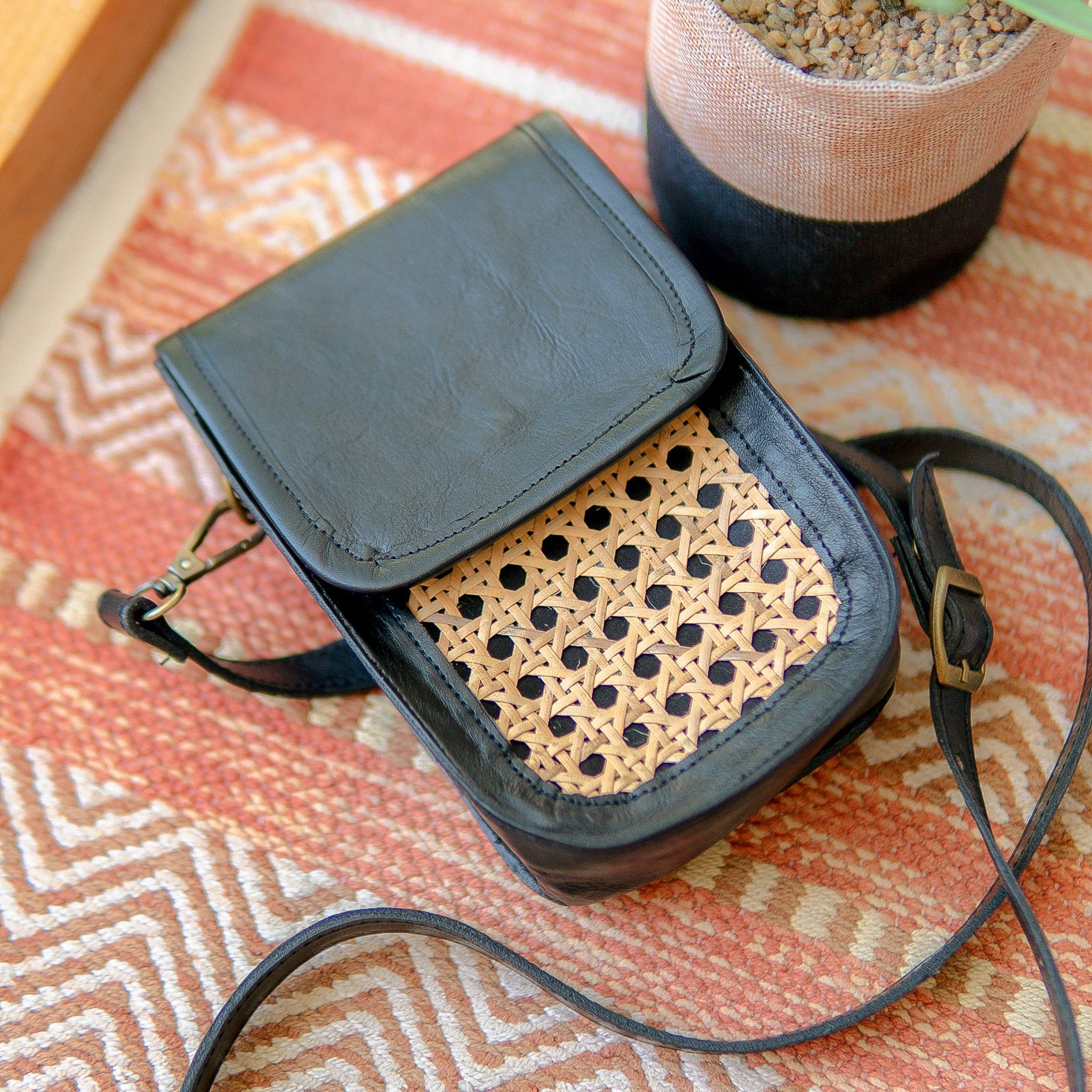Black leather crossbody bag with woven texture on a patterned surface. Harper Sling Bag in black leather with rattan weave