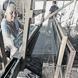A woman artisan weaving a handloom blanket using a traditional wooden loom in a rustic workshop. She carefully threads the fabric, showcasing the intricate craftsmanship involved in creating handwoven textiles. Natural light filters through the brick lattice wall, highlighting the artisan’s dedication to preserving traditional weaving techniques.