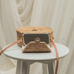 Woven handbag with leather strap on a small round table against a white curtain background