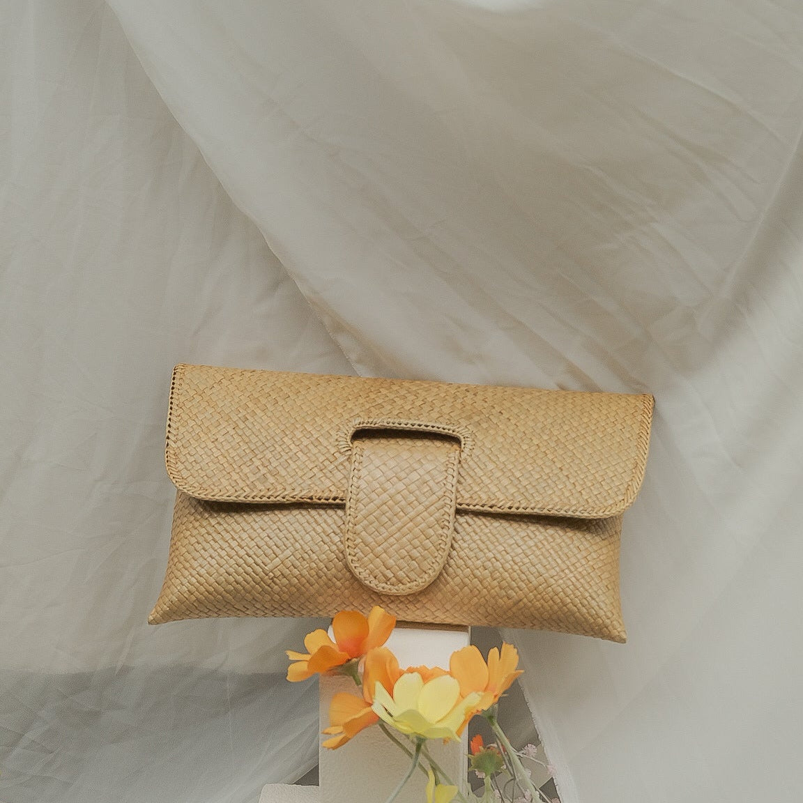 Beige woven clutch bag with a small bouquet of flowers on a white fabric background