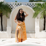 Woman in an orange dress standing by a pool with palm trees and a white archway in the background.