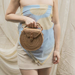 Woman holding a woven handbag wearing a strapless dress with a blue and beige pattern.