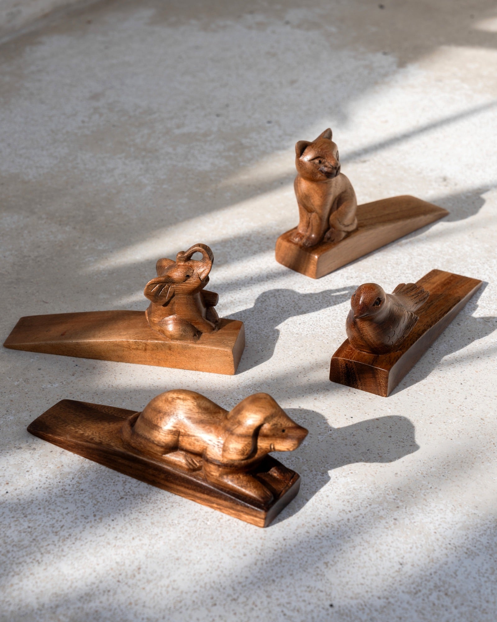 Four small carved wooden animal figurines displayed on rectangular wooden bases against a light concrete wall, with natural sunlight casting shadows.