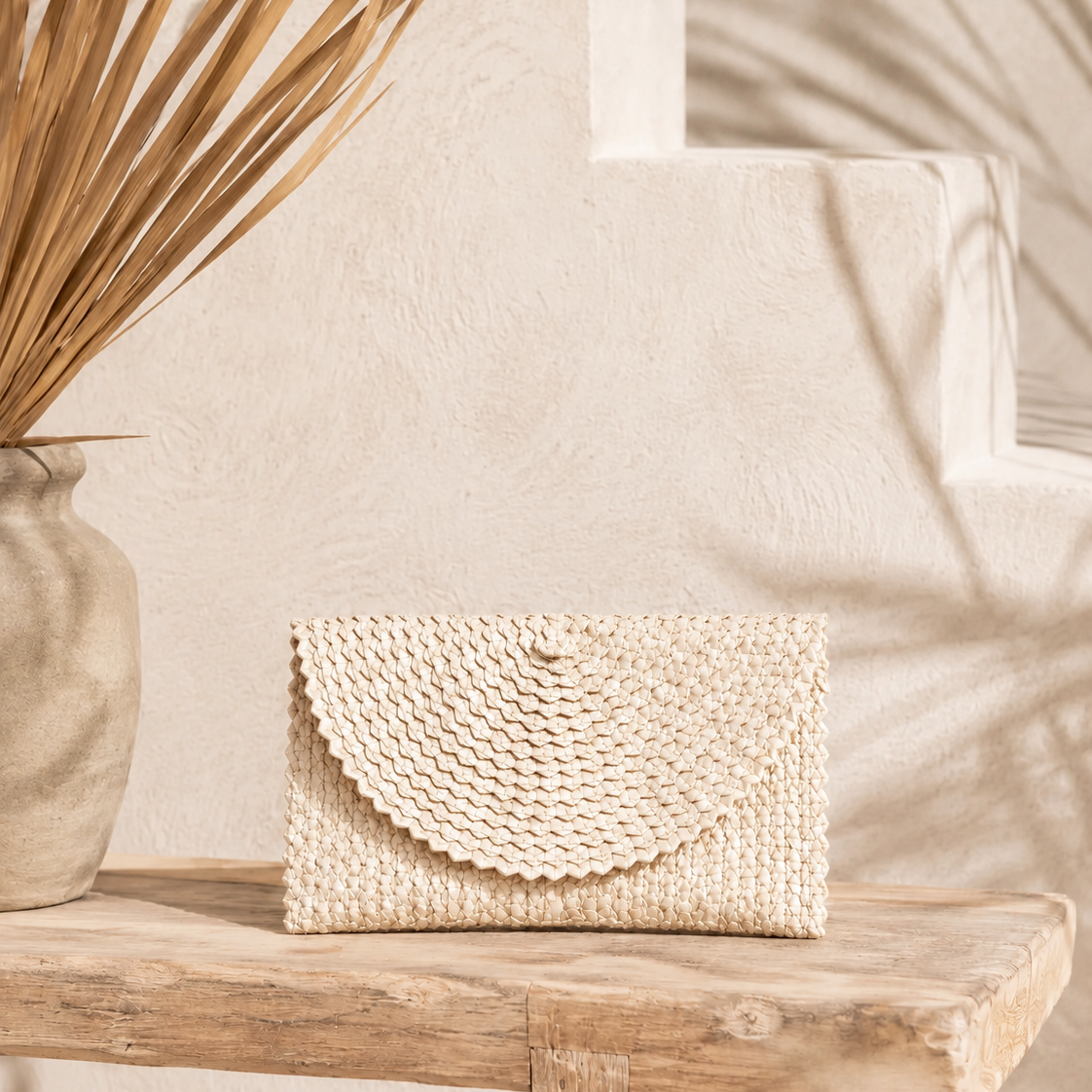 Aya Palm Leaf Clutch in natural cream, hand woven with scalloped flap, displayed on a rustic timber bench.