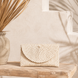 Aya Palm Leaf Clutch in natural cream, hand woven with scalloped flap, displayed on a rustic timber bench.
