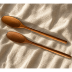 Two wooden spoons with a warm medium-brown finish, arranged diagonally on a textured beige fabric background under soft, warm sunlight.