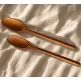 Two wooden spoons with a warm medium-brown finish, arranged diagonally on a textured beige fabric background under soft, warm sunlight.