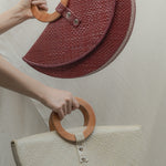 Two half-circle handbags with wooden handles held by a person against a neutral background.