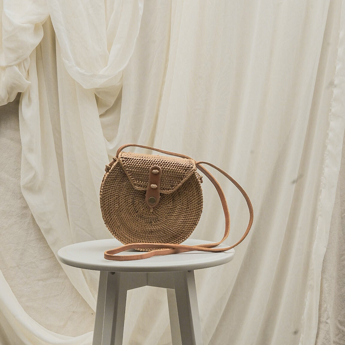 Woven handbag with leather strap on a small white stool against a draped white curtain background
