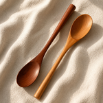 Two wooden spoons, one dark brown and one light honey brown, arranged diagonally on a textured beige fabric background with soft shadows.