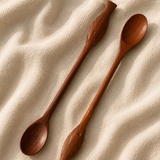 Two long wooden spoons with decorative carved handles, arranged diagonally on a textured beige fabric background.