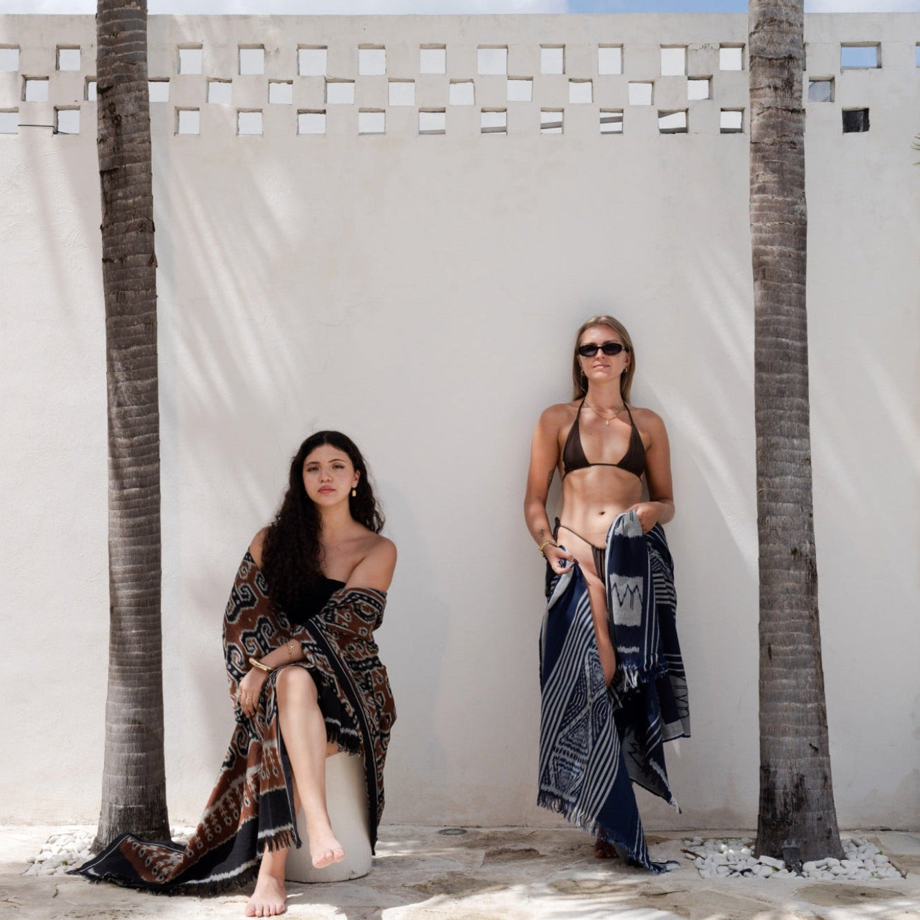 Two women posing outdoors against a white wall with palm trees. One woman sits on a stone block wearing a dark patterned wrap dress, and the other stands wearing a black bikini top with a matching dark patterned sarong.