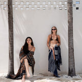 Two women posing outdoors against a white wall with palm trees. One woman sits on a stone block wearing a dark patterned wrap dress, and the other stands wearing a black bikini top with a matching dark patterned sarong.