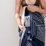 A woman wearing a dark brown halter bikini top and holding a navy blue and white patterned silk scarf wrapped around her lower body, standing against a plain light gray wall.