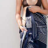 A woman wearing a dark brown halter bikini top and holding a navy blue and white patterned silk scarf wrapped around her lower body, standing against a plain light gray wall.