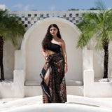 A woman stands in front of a white arched wall and pool, wearing a black one-shoulder top and a long black maxi skirt with a brown and beige patterned print. Palm trees and a blue sky are visible in the background.