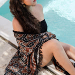 A woman sitting by a swimming pool wearing a brown, black, and beige patterned wrap skirt with fringe details.