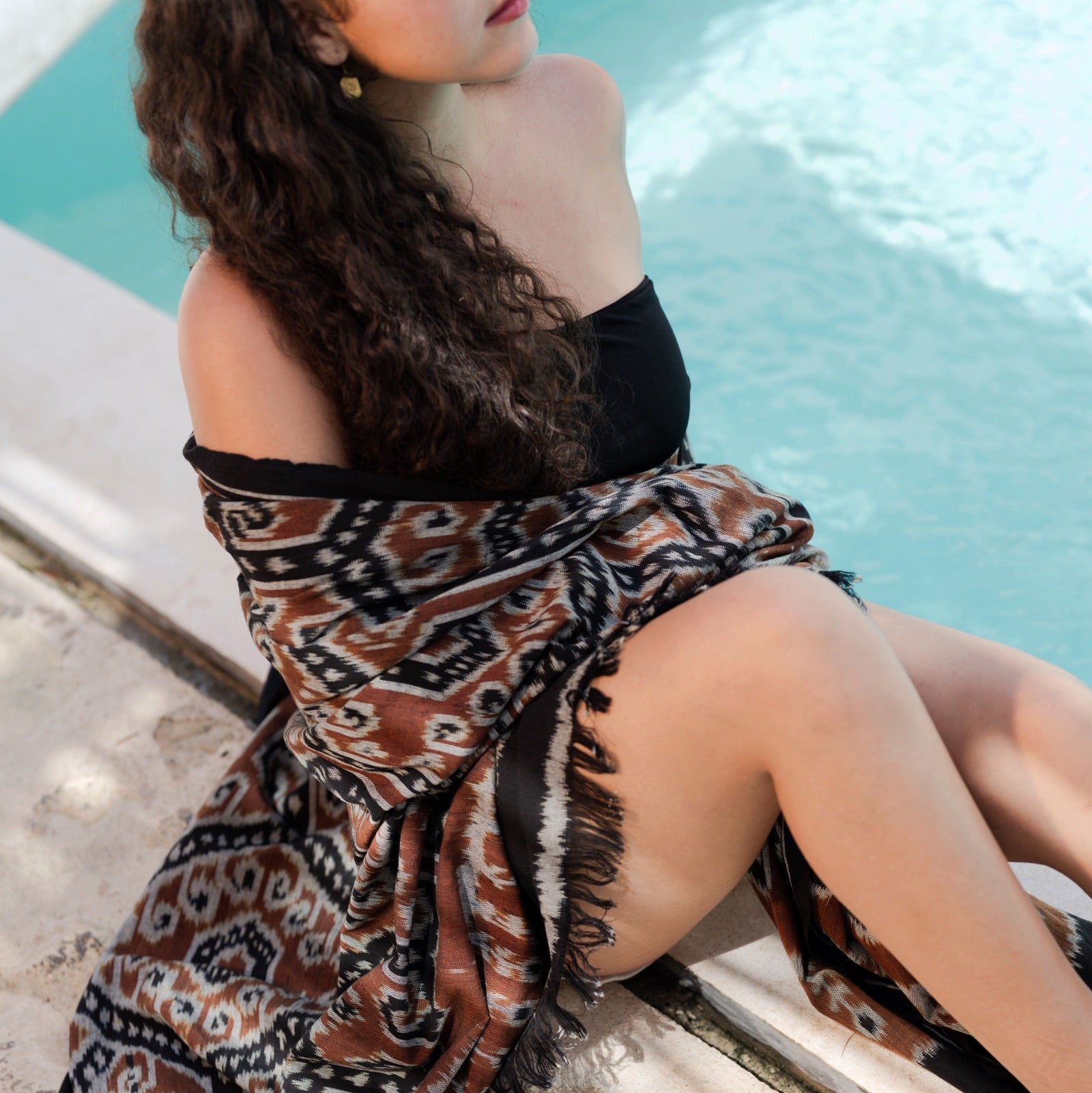 A woman sitting by a swimming pool wearing a brown, black, and beige patterned wrap skirt with fringe details.