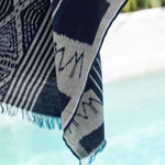 A close-up of a woven throw blanket with a navy blue and light gray ikat pattern, featuring fringed edges, draped over a white poolside surface with a blurred outdoor background.