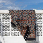 A patterned textile runner or throw draped over a white decorative wall under a blue sky, with a white cushion in the foreground.