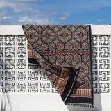 A patterned textile runner or throw draped over a white decorative wall under a blue sky, with a white cushion in the foreground.