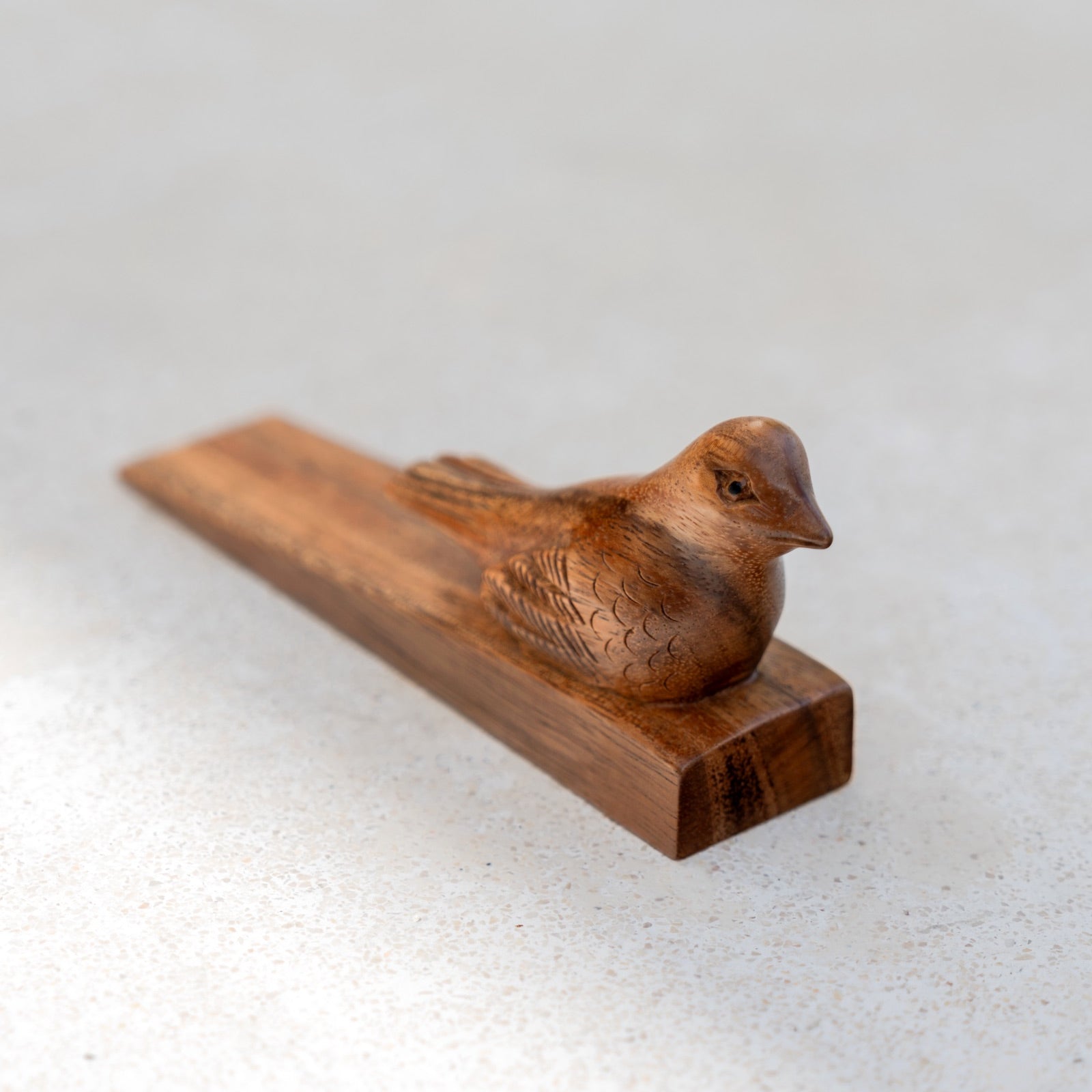 A small carved wooden bird figurine resting on a rectangular wooden base, photographed on a light neutral textured surface.