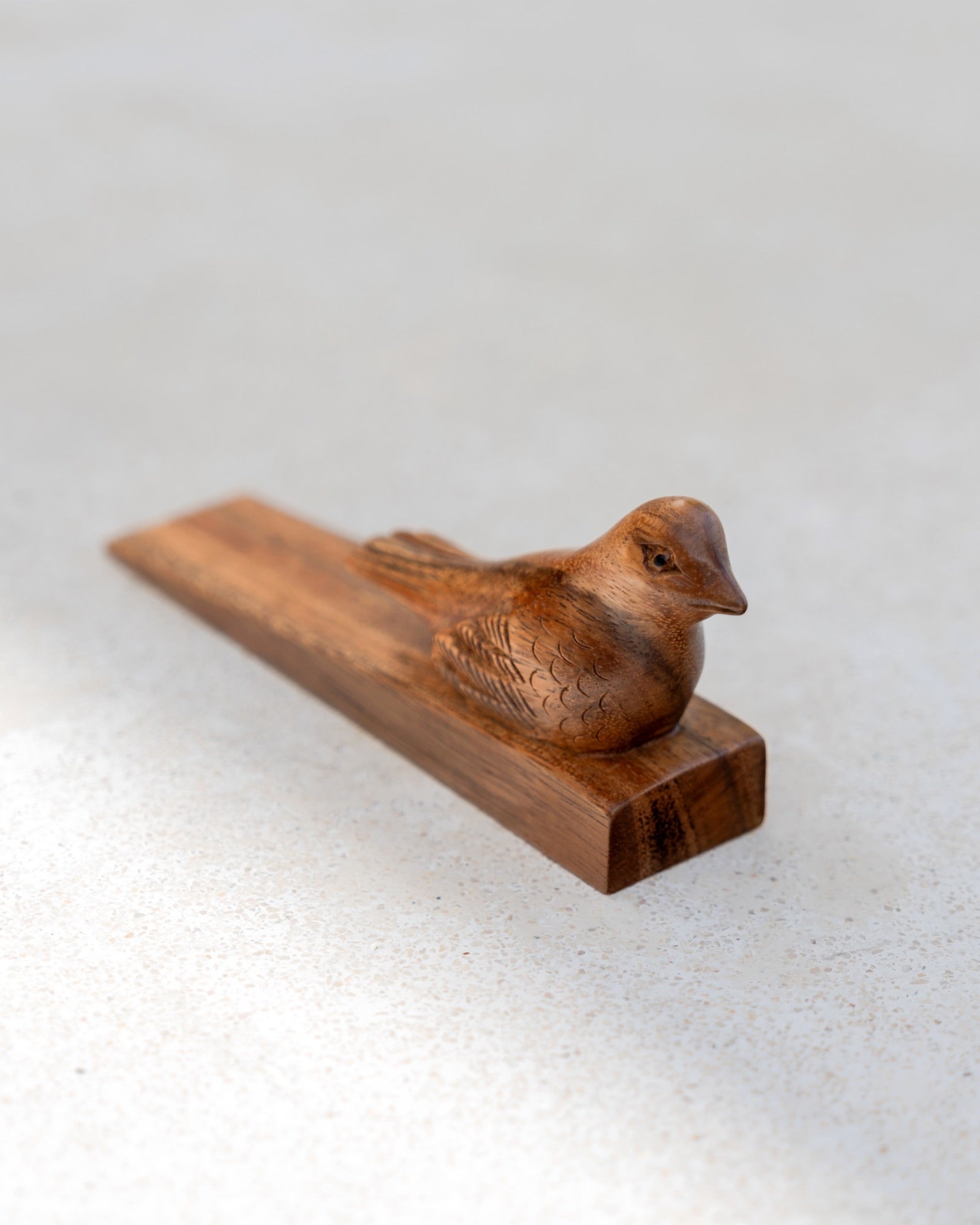 A small carved wooden bird figurine resting on a rectangular wooden base, photographed on a light neutral textured surface.