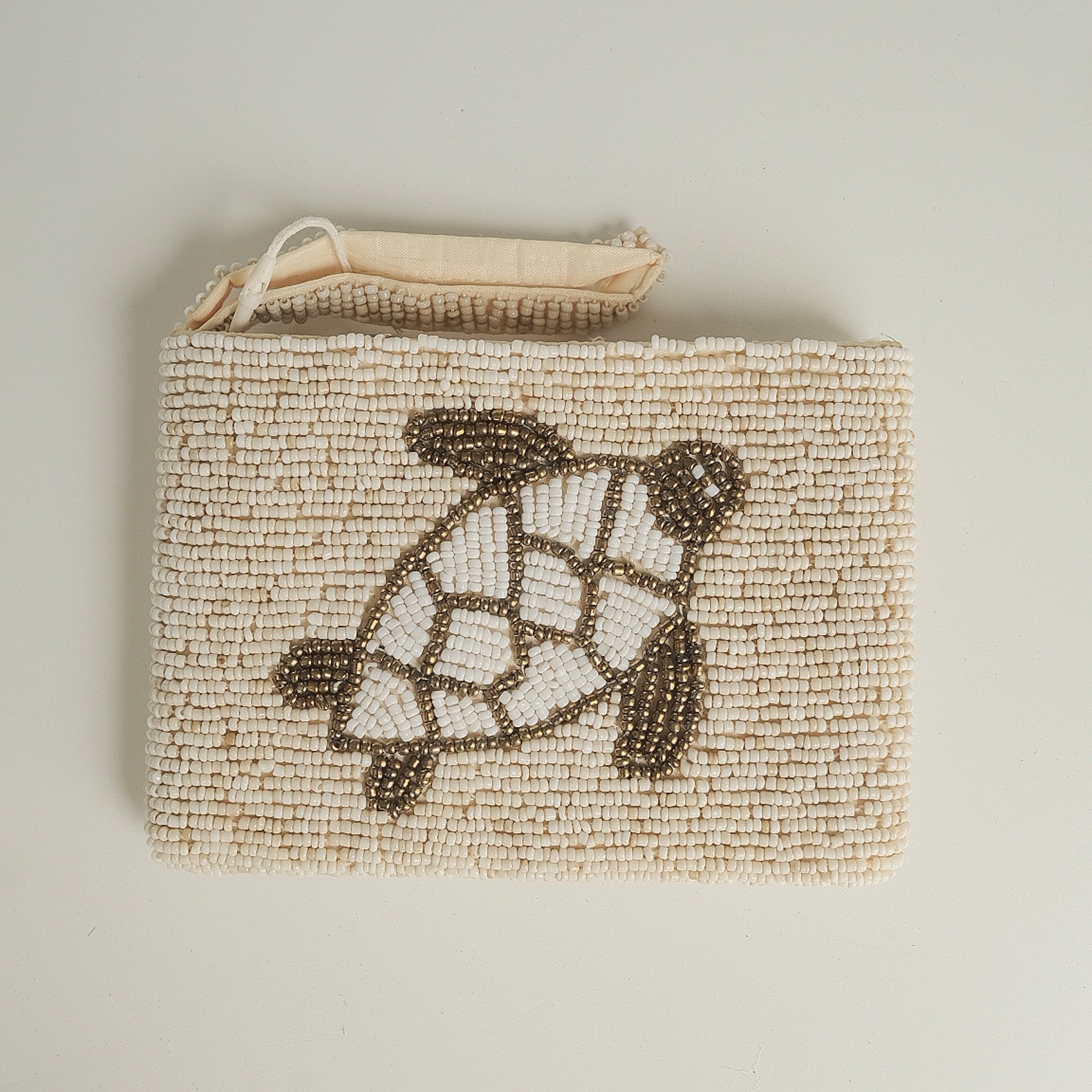 A square woven clutch bag in natural beige with a beaded turtle design in cream and brown, featuring a top zipper closure, photographed on a neutral light gray background.