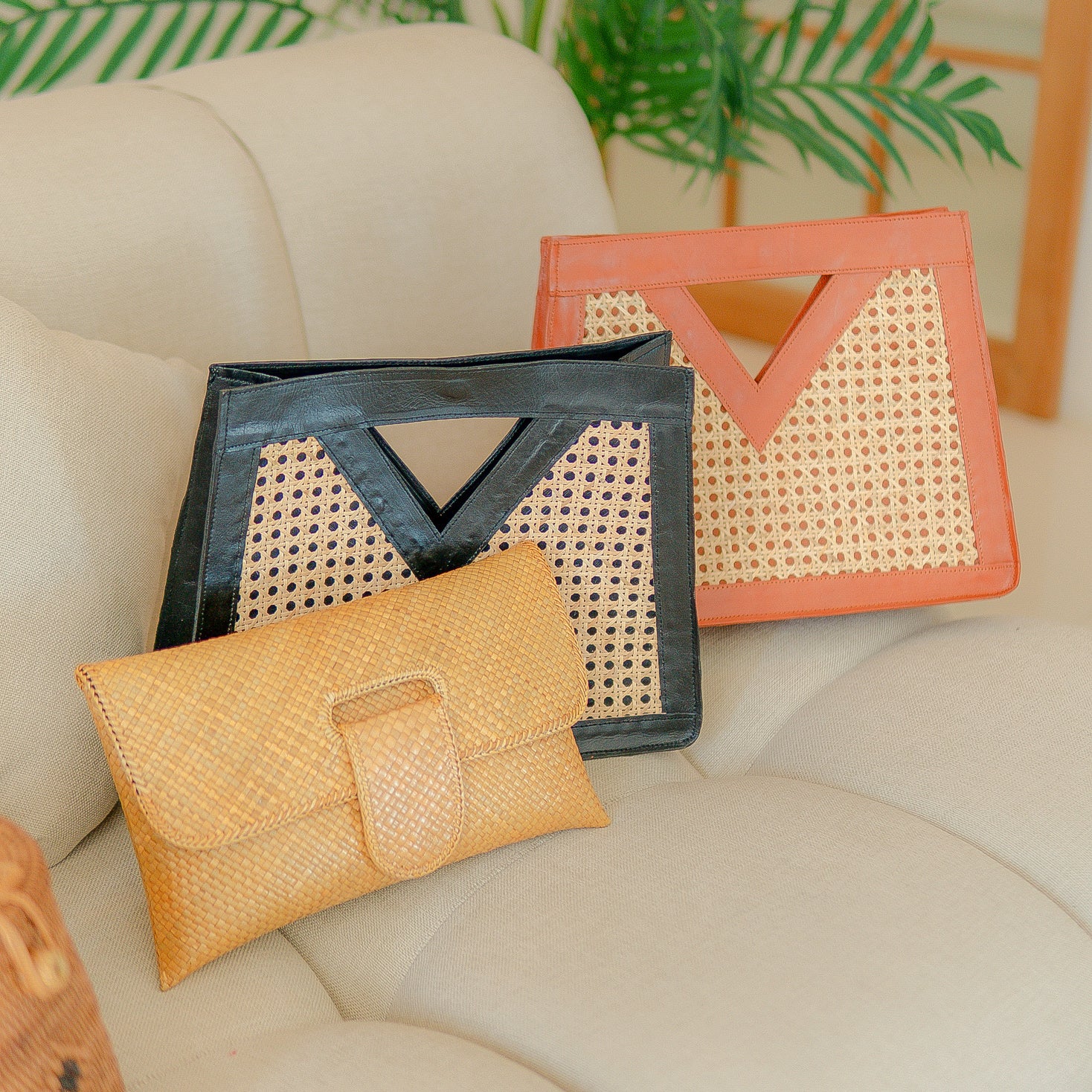 Three woven handbags in black, orange, and yellow on a beige couch with green plants in the background.