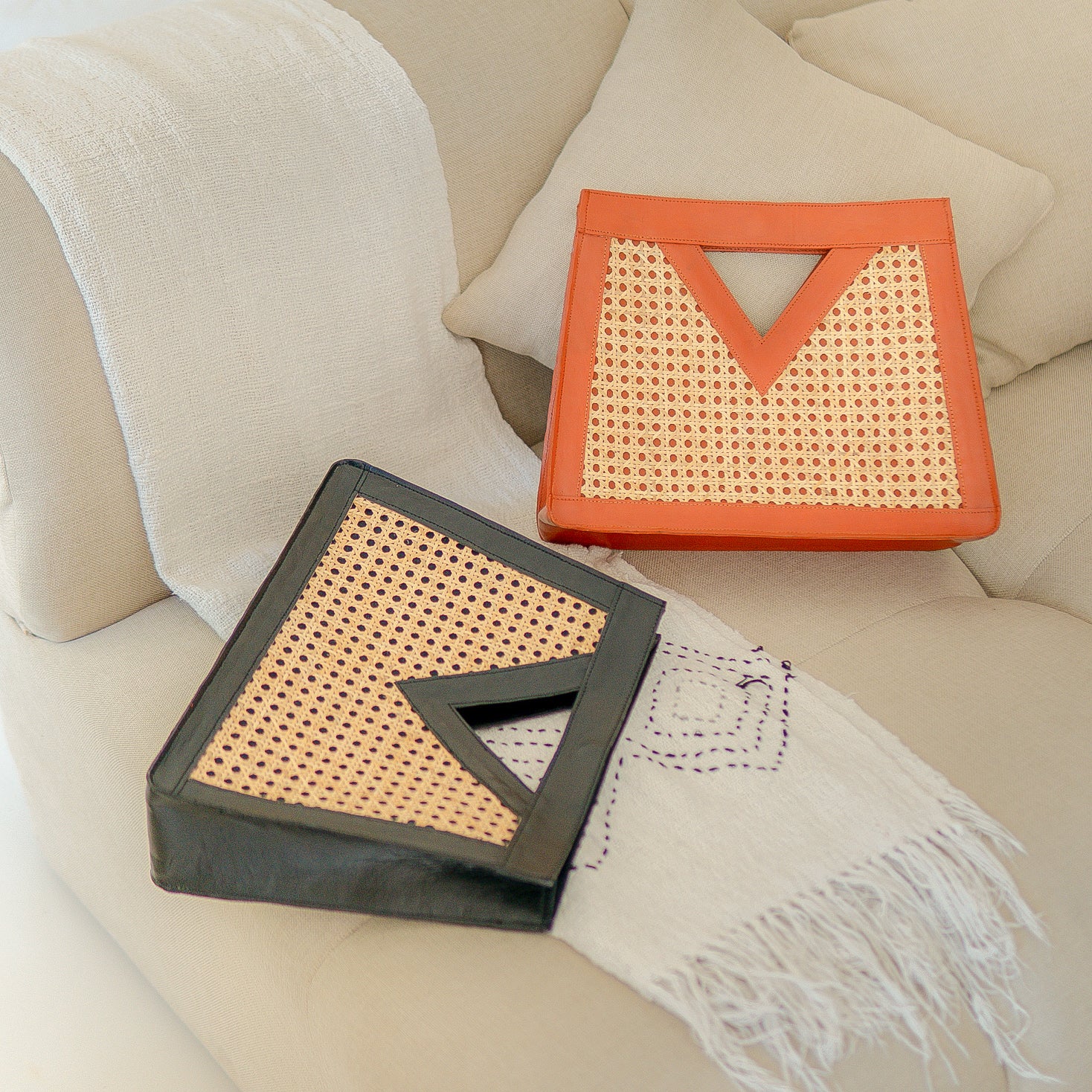 Two rattan woven boxes, one black leather and one tan leather, on a beige sofa with a white blanket.