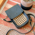 Black leather crossbody bag with woven texture on a patterned surface. Harper Sling Bag in black leather with rattan weave