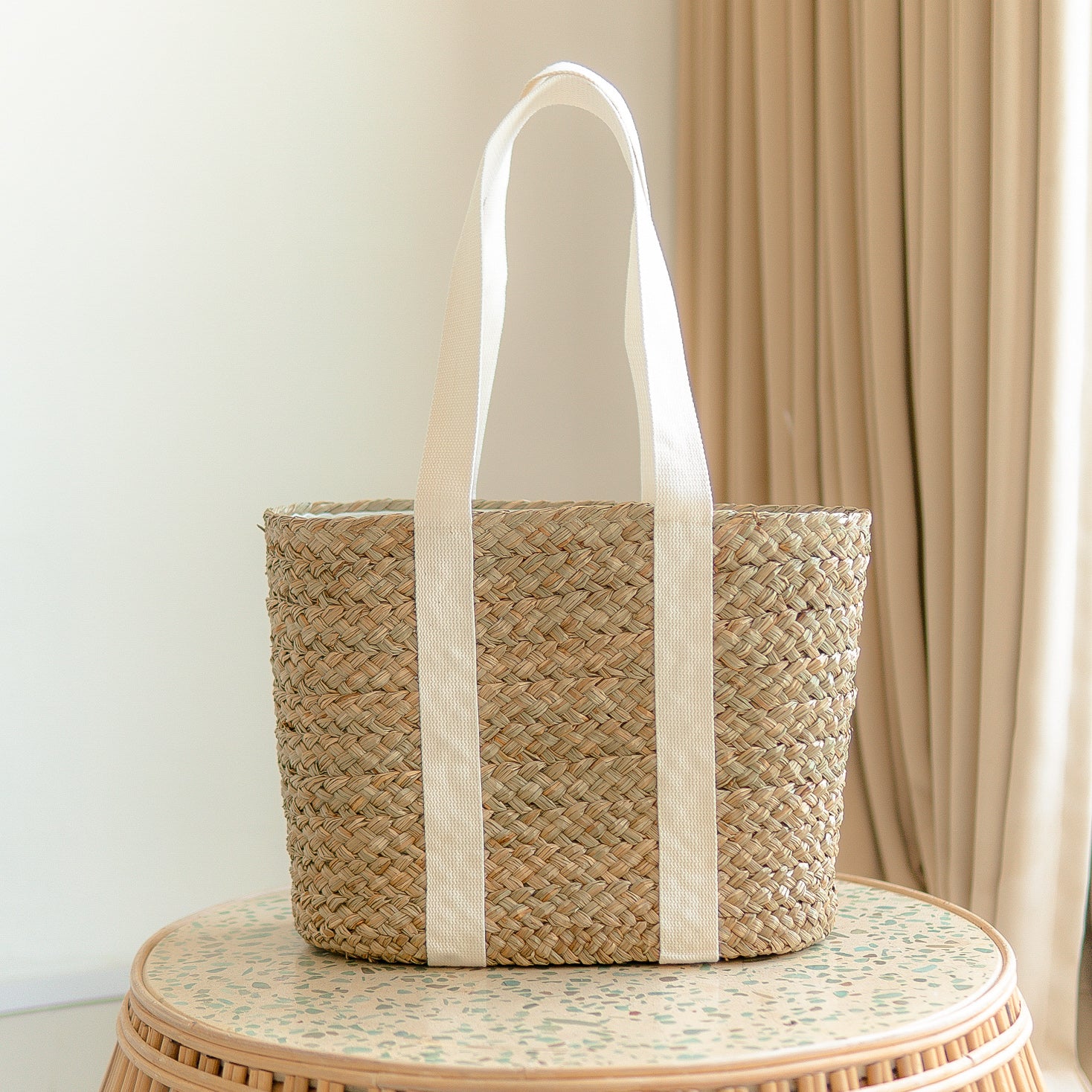 Isla Tote Bag – handwoven water hyacinth with white cotton straps. Woven basket with white handle on a round table against a beige curtain background