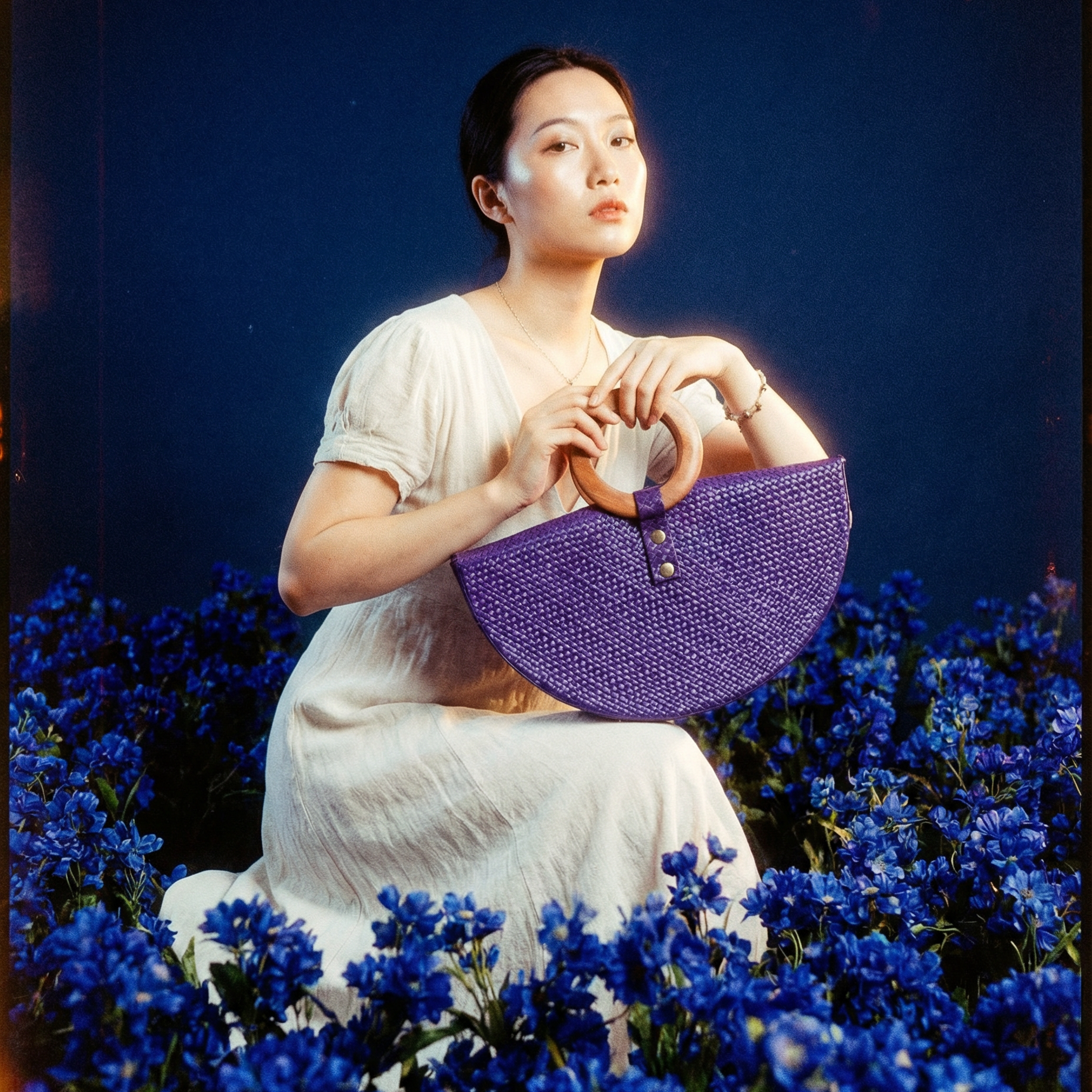 Woman holding a purple woven handbag in a field of blue flowers with a dark blue background