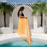 Woman in a yellow dress standing by a pool with palm trees and a white architectural structure in the background.