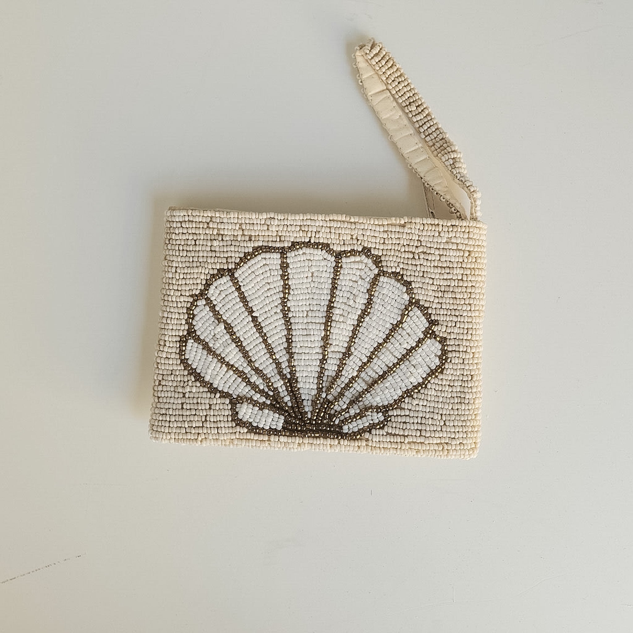A small rectangular beaded clutch or wristlet with a shell motif on the front, featuring a cream-colored woven strap, photographed against a plain off-white background.